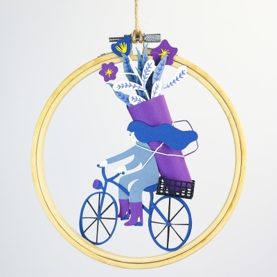 Decoração em anel de bordado com papel recortado de mulher numa bicicleta azul carregando flores