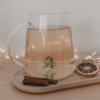 Chaleira de vidro com líquido âmbar, pinheiro verde no fundo, paus de canela e rodelas de fruto seco em tabuleiro de madeira