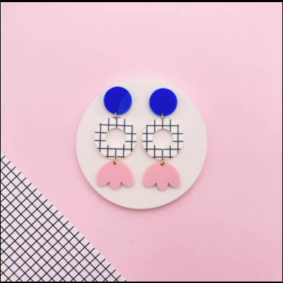 Brincos coloridos com formas geométricas sobre fundo rosa e padrão de grelha