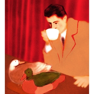 Homem com casaco bege a beber chávena com dois patos empalhados numa mesa.