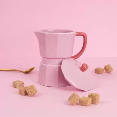 Cafeteira moka rosa com pega salmão, tampa, cubos de açúcar e colher dourada sobre fundo rosa.