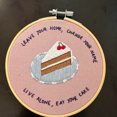 Bordado de fatia de bolo num bastidor de madeira com texto em inglês em fundo rosa