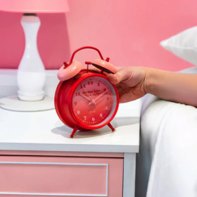 Relógio despertador analógico vermelho com sino rosa numa mesa de cabeceira branca e rosa ao lado de uma mão e candeeiro branco