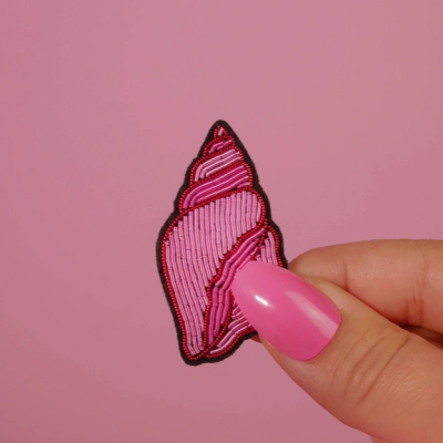 Patch bordado em forma de concha rosa segurado por mão com unha pintada de rosa