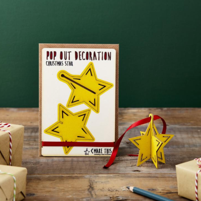 Caixa branca com decoração pop out Christmas star amarela e fita vermelha numa mesa de madeira.