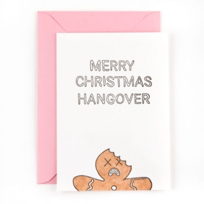 Cartão de Natal branco com envelope rosa, texto 'MERRY CHRISTMAS HANGOVER' e biscoito de gengibre triste mordido.