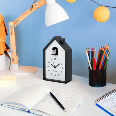 Relógio de mesa formato casa preto e branco com lâmpada, lápis coloridos e cadernos numa mesa branca