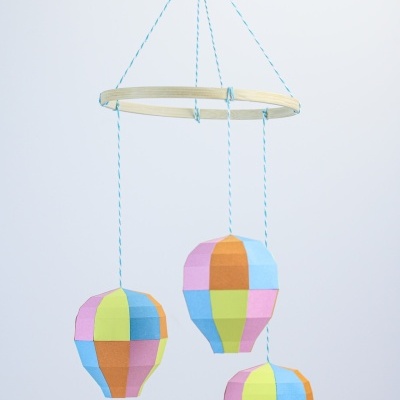 Mobile decorativo com três balões coloridos suspensos por fios azuis e brancos numa estrutura circular de madeira