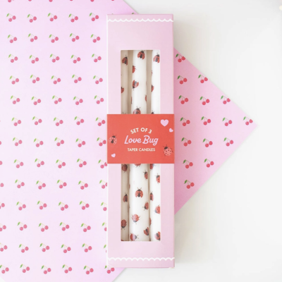 Conjunto de 3 velas cónicas decoradas com padrão de joaninhas na caixa rosa com etiqueta vermelha sobre papel decorado com padrão de cerejas