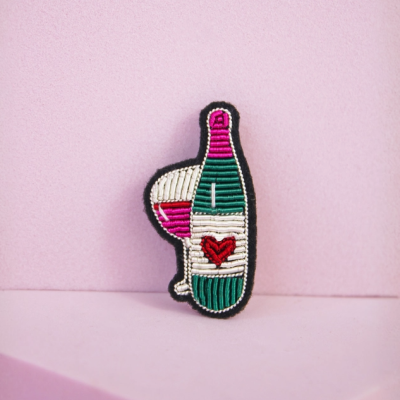 Broche bordado em forma de garrafa de vinho e copo em fundo rosa.