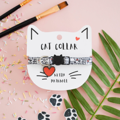 Colar para gato com padrão e pendentes em cartão branco em forma de rosto de gato sobre fundo rosa com folhas e pincéis