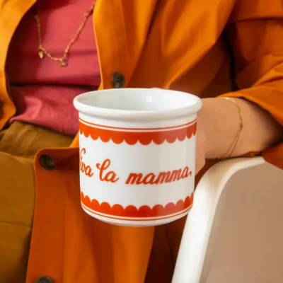 Caneca branca com padrão e texto vermelho, segurada por pessoa com casaco laranja.