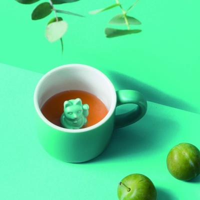 Caneca verde com infusor de chá em forma de gato e duas ameixas verdes