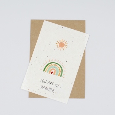 Cartão branco reciclado com sol, arco-íris e texto 'YOU ARE MY SUNSHINE' sobre envelope castanho