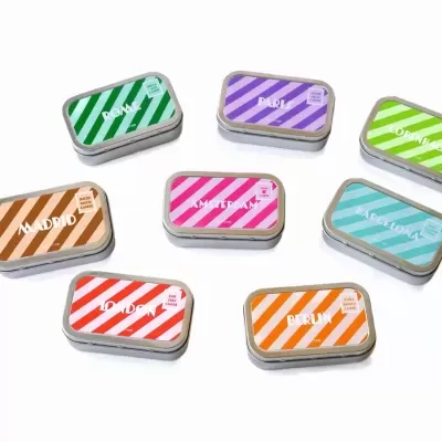 Sete latas retangulares pequenas com tampas coloridas listradas sobre fundo branco