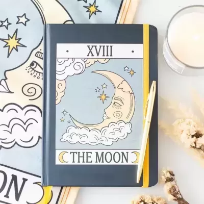 Caderno preto com ilustração da lua, caneta dourada, vela e flores secas numa mesa clara.