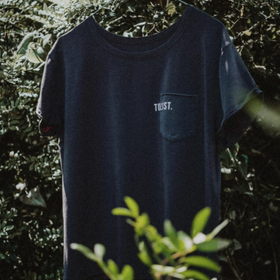 T-shirt azul escura com bolso e texto TOUST pendurada entre folhas verdes