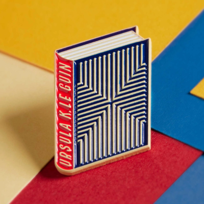 Broche de livro azul e branco com lombada vermelha e texto branco URSULA K. LE GUIN