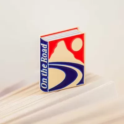 Pin metálico em forma de livro com design de estrada e texto 'On the Road'