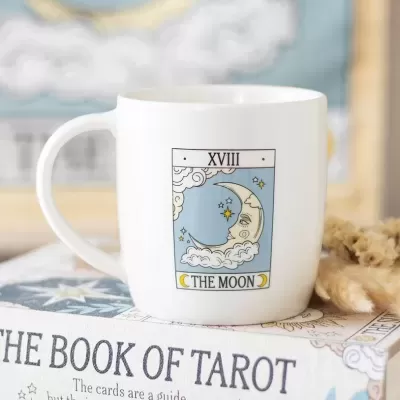 Caneca branca com ilustração do tarot The Moon sobre livro The Book of Tarot