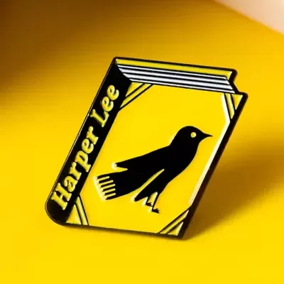 Pino em forma de livro amarelo com texto 'Harper Lee' e imagem de pássaro preto