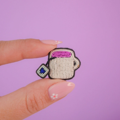 Pin bordado em forma de caneca de chá com saqueta, segurado por dedos com unhas decoradas e fundo lilás