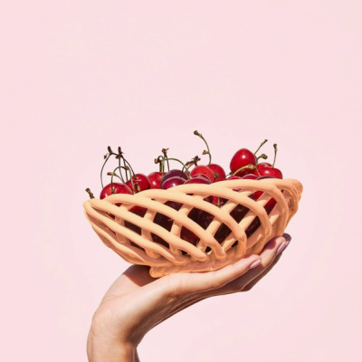 Cesto entrelaçado salmão cheio de cerejas vermelhas segurado por uma mão com fundo rosa claro