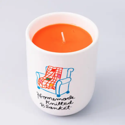 Vela laranja em copo branco decorado com desenho de poltrona e manta