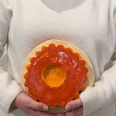 Peça cerâmica em forma de rosca laranja e bege, segurada por pessoa com suéter branco.