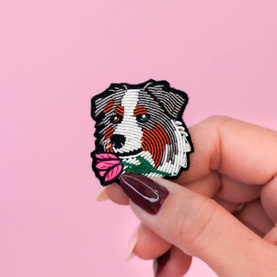 Broche bordado de cão segurando flor numa mão com fundo rosa.