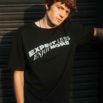 Homem usando t-shirt preta com frases em branco 'EXPECT LESS ENJOY MORE'