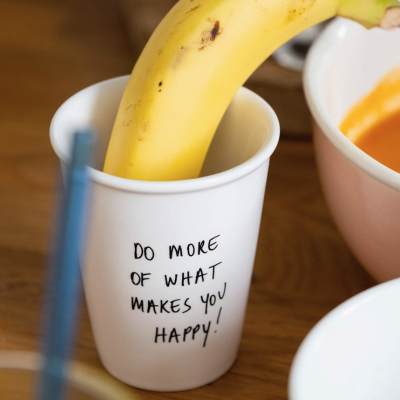 Copo branco com texto motivacional e banana dentro numa mesa de madeira