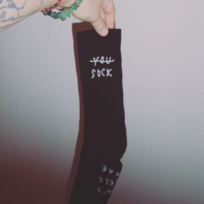 Meia preta com texto branco 'YOU SOCK' segurada por mão tatuada com pulseiras.