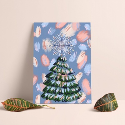 Cartão de Natal com árvore verde e estrelas, fundo azul com manchas coloridas, sobre superfície clara com folhas secas