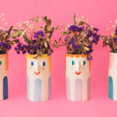 Quatro vasos de cerâmica com rostos pintados e flores secas sobre fundo rosa