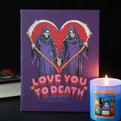 Quadro roxo com esqueletos e texto, vela acesa com etiqueta colorida numa mesa preta