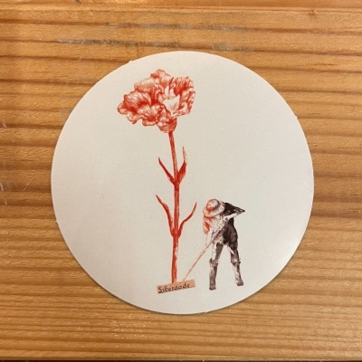 Autocolante redondo com desenho de flor vermelha e figura humana junto à palavra 'Liberdade' sobre fundo de madeira.