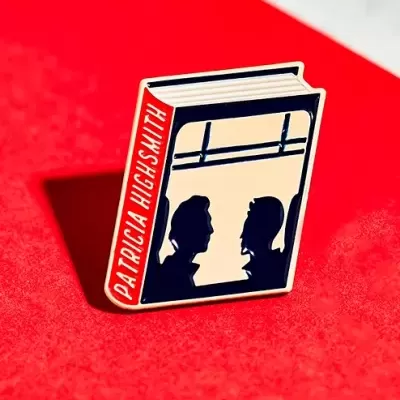 Alfinete esmaltado em forma de livro com silhuetas humanas em capa e texto 'PATRICIA HUGHESMITH' na lombada.