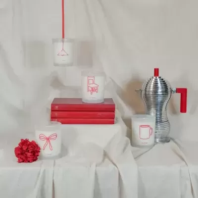 Velas em vidro fosco com desenhos vermelhos e jarro metálico com pega vermelha sobre tecido branco.