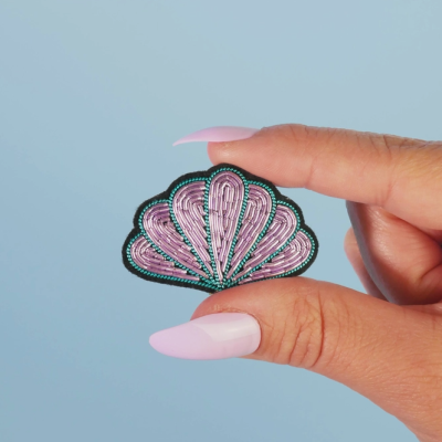 Patch bordado em forma de concha cor de rosa e verde, segurado por dedos com unhas compridas cor de rosa.