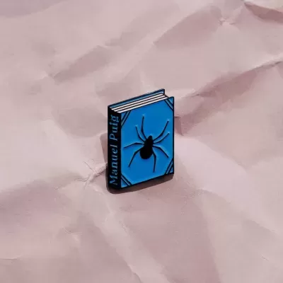 Pin azul em forma de livro com aranha preta sobre fundo de papel cor-de-rosa amassado