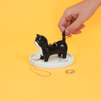 Suporte de joias cerâmico em forma de gato preto e branco com anéis e colar numa base branca sobre fundo amarelo