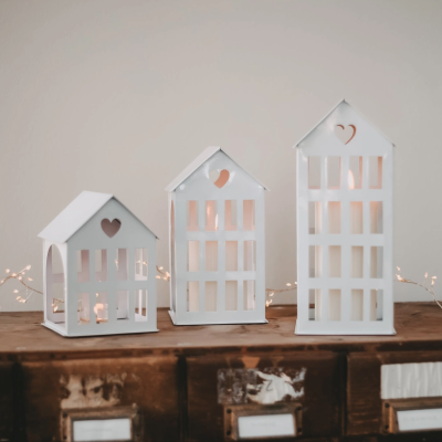 Três lanternas brancas em forma de casa sobre móvel de madeira com luzes acesas