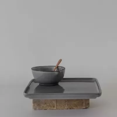 Taça cinzenta de cerâmica com colher de madeira numa bandeja cinzenta apoiada em bloco de pedra.