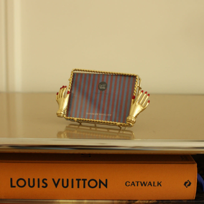 Porta-foto dourado com mãos decorativas e tecido listrado, sobre livros LOUIS VUITTON e JIMMY NELSON