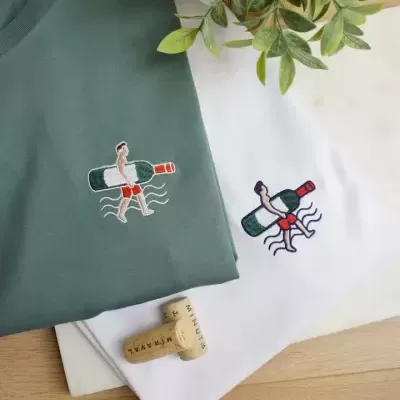 Dois t-shirts, uma verde escura e outra branca, com bordado de figura humana com garrafa de vinho verde, três rolhas e planta