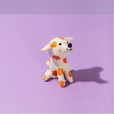 Figura decorativa de cão em cerâmica branca com manchas laranja sobre fundo lavanda