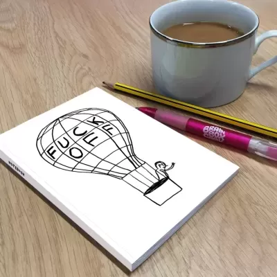 Bloco de notas com desenho de balão de ar quente e texto F*** OFF, lápis, caneta rosa e caneca com bebida sobre mesa de madeira.