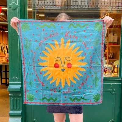 Lenço azul com sol amarelo e texto vermelho, segurado em frente a uma loja verde