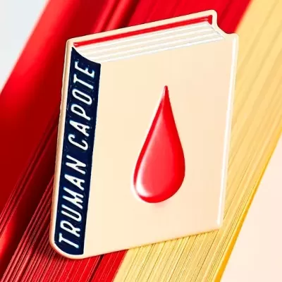 Broche em forma de livro com texto TRUMAN CAPOTE e uma gota vermelha, sobre páginas de livros vermelhas e amarelas